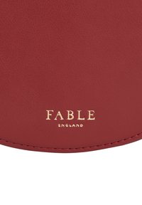 Rød læderoverflade med en tekstureret finish, prydet med præget guldtekst "FABLE" og "ENGLAND" langs bunden.