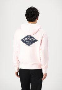 Rosa Kapuzenpullover mit Kordelzug, ausgestattet mit einem dunkelblauen Diamanten-Logo, das auf dem Rücken "TOMMY JEANS" aufgedruckt ist, und Rippbündchen sowie -saum.