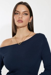 Top off-shoulder a coste di colore blu navy con maniche lunghe, realizzato in un tessuto testurizzato e adornato da una collana in oro con un pendente decorativo.