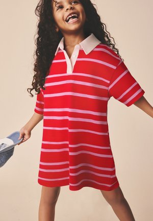 Niño sonriente con cabello largo y rizado, vestido con un vestido de rayas rojas y rosas, sosteniendo un sombrero gris en una mano, de pie frente a un fondo beige.