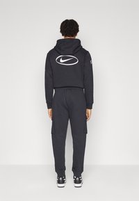 Svart Nike-hoodie med logotyp på baksidan, ihop med matchande svarta cargobyxor. Har en avslappnad passform och dragsko i huvan. Svart- och vit sneakers.