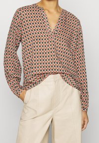 Blouse à manches longues avec un col en V, présentant un motif géométrique rouge, vert et blanc sur un tissu léger. Associée à un pantalon beige.