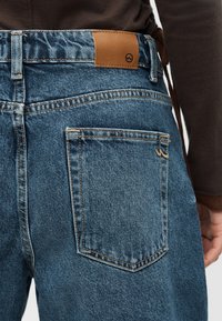 Πίσω όψη ατόμου που φοράει μπλε τζιν από denim με δερμάτινη ετικέτα και μία πίσω τσέπη, ντυμένο με σκούρο μακρυμάνικο πάνω μέρος.