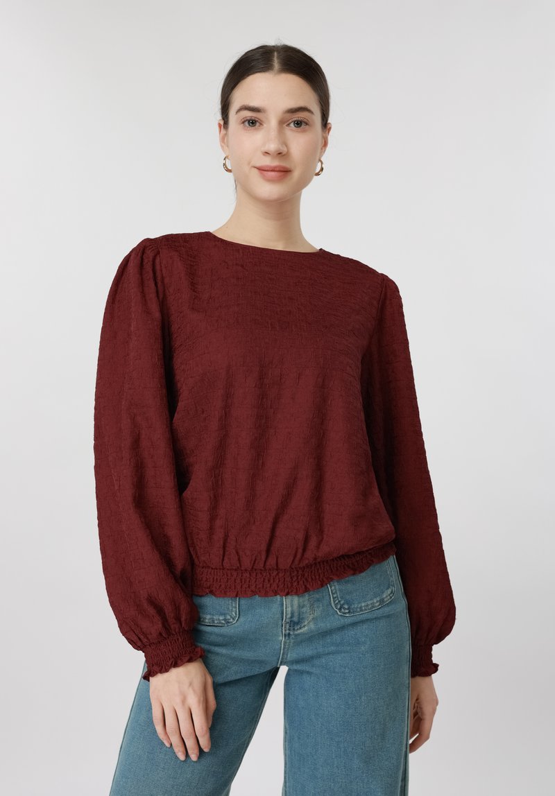 Γυναίκα που φοράει μπλούζα μπορντό με υφή, μακρυμάνικη με φουσκωτά μανίκια και λάστιχο στα μανίκια, σε συνδυασμό με μπλε τζιν, πάνω σε απλό φόντο.