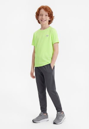 Băiat cu părul roșcat zâmbind, stând cu mâinile în buzunare, purtând un tricou verde neon, pantaloni gri închis și adidași gri pe fundal alb.