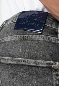 Tmavě šedé džíny z denimu mají lesklou námořnickou koženou nášivku s reliéfním nápisem "SCOTCH & SODA AMSTERDAM". Viditelné stehy podél zadních kapes.