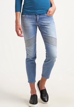 Persoon draagt lichtblauwe slim-fit jeans met geribbelde details op de knieën, blauwe top met lange mouwen en zwarte instapschoenen met witte zolen.