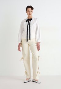 Mujer vistiendo una blusa blanca de manga larga con corbata negra y pantalones crema con lazos laterales, combinados con bailarinas plateadas, de pie contra una pared blanca.