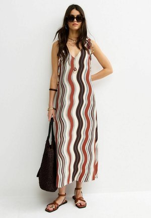 Femme portant une robe maxi sans manches à rayures ondulées en marron, rouille et crème, tenant un sac fourre-tout tressé noir, et portant des lunettes de soleil et des sandales.