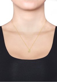 Guld kæderekæde med en rund vedhæng, der har et sammenflettet design. Kæden er sart og sidder lige over kravebenet.