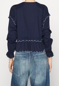 Blusa blu navy con orlo a strati e bordo ondulato, abbinata a jeans in denim blu sbiadito a vita alta con tasche posteriori.