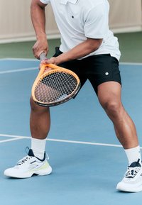 Raquete de ténis com estrutura laranja e pega preta, segurada por uma pessoa vestindo uma polo branca e calções pretos, em pé num campo azul com linhas brancas.