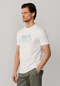 Mann trägt ein weißes Kurzarm-T-Shirt mit blauem Palmenmotiv und Text, kombiniert mit grüner Hose, steht vor einem einfarbigen grauen Hintergrund.