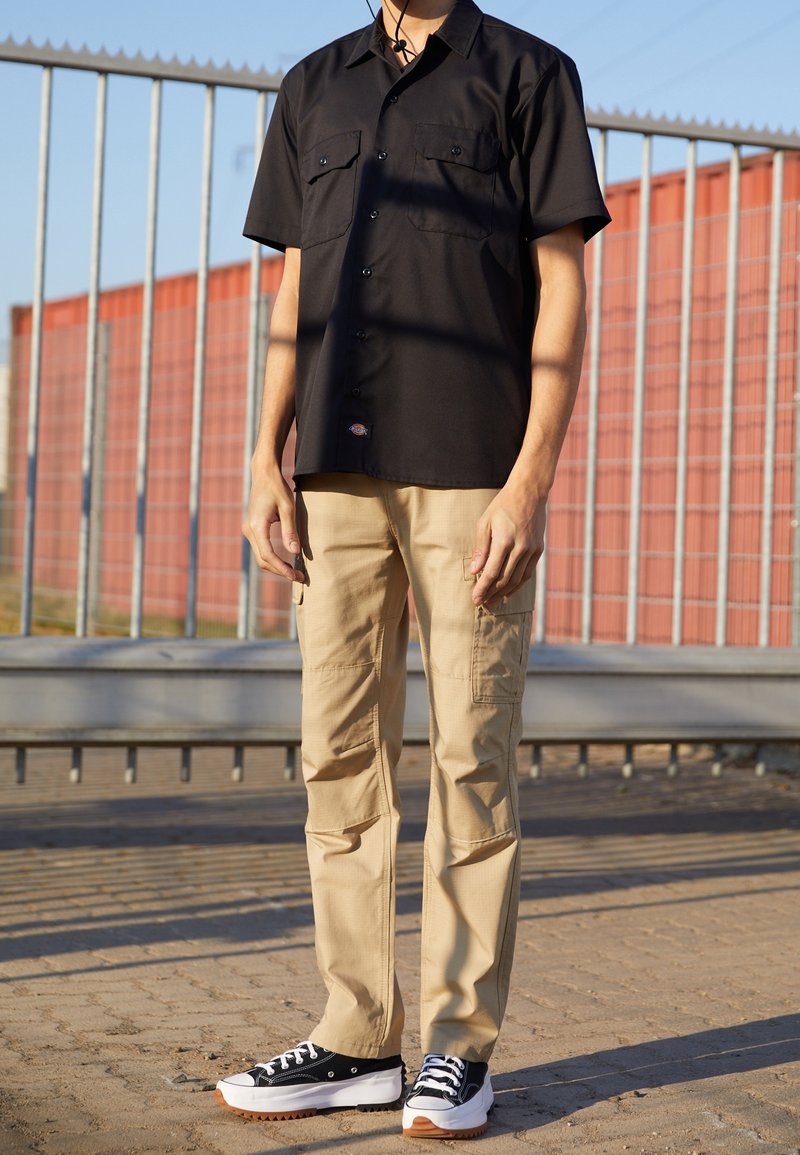 Schwarzes Kurzarmhemd mit zwei Brusttaschen, kombiniert mit beigen Cargo-Hosen und schwarzen Sneakers mit weißen Sohlen und braunen Akzenten.
