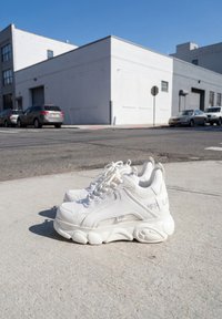 Par hvide chunky sneakers med teksturerede såler placeret på en betonfortov under en klar himmel nær et gadehjørne med parkerede biler.