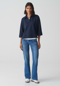 Pull bleu marine avec col, manches larges, superposé sur une chemise blanche. Associé à un jean évasé bleu clair et des baskets blanches.