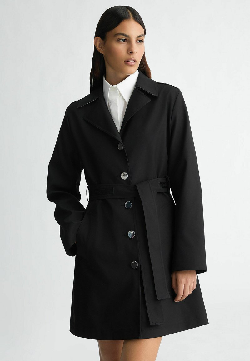 Cappotto trench nero con collo a bavero, chiusura con bottoni e cintura in vita. Tessuto morbido con una forma classica e aderente.