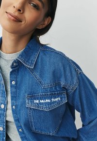 Denim jacket in blue with a button-up front, pointed collar, and a chest pocket featuring "The Rolling Stones" printed in white.