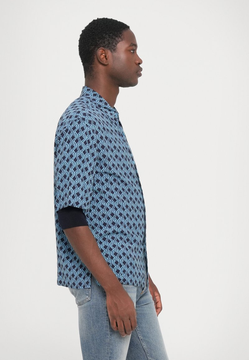 Joven enfrentando a la derecha, vistiendo una camisa de manga corta azul con un patrón sobre una camisa de manga larga negra y jeans azul claro, contra un fondo blanco.