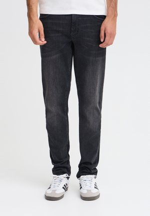 Un homme porte un jean noir à coupe droite, un tee-shirt blanc et des baskets blanches sur un fond de studio uni.