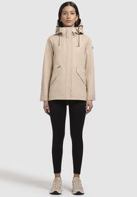 Beige waterdichte jas met een hoge kraag, verstelbare capuchon, twee voorzakken en een ritssluiting aan de voorkant, gecombineerd met zwarte leggings en sneakers.