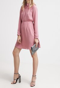 Lyserød kjole lavet af let stof, med krave, blonder og elastisk talje. Sammen med sølv høje hæle og en clutch.
