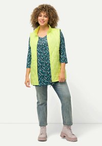 Gilet trapuntato verde lime con colletto alto sopra una maglietta floreale teal a maniche lunghe, abbinato a jeans sbiaditi e stivali chunky rosa chiaro.