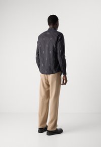 Camicia a righe nere con motivi bianchi, abbinata a pantaloni beige e scarpe nere, mostrata da una vista posteriore.
