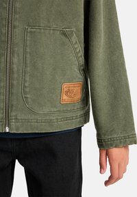 A close-up of a person's hand holding the zipper of a green jacket, featuring a brown branded patch on the front pocket.