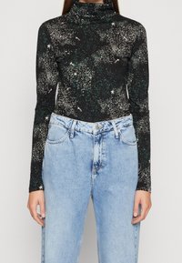 Svart långärmad polotröja med ett stjärnmönster, som har vita och gröna prickar. Kombineras med ljusblå högmidjade jeans.