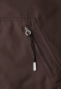 Gros plan sur une poche en tissu marron avec une fermeture éclair argentée et des cordons noirs se terminant par des embouts blancs.