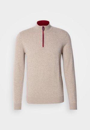 Suéter beige de cuello alto con una cremallera corta. Presenta detalles rojos a lo largo de la cremallera y una textura suave y lisa.