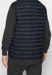 Padded navy vest with vertical quilting, shiny finish, and sleeveless design. Textured fabric and elastic waistband for comfort.