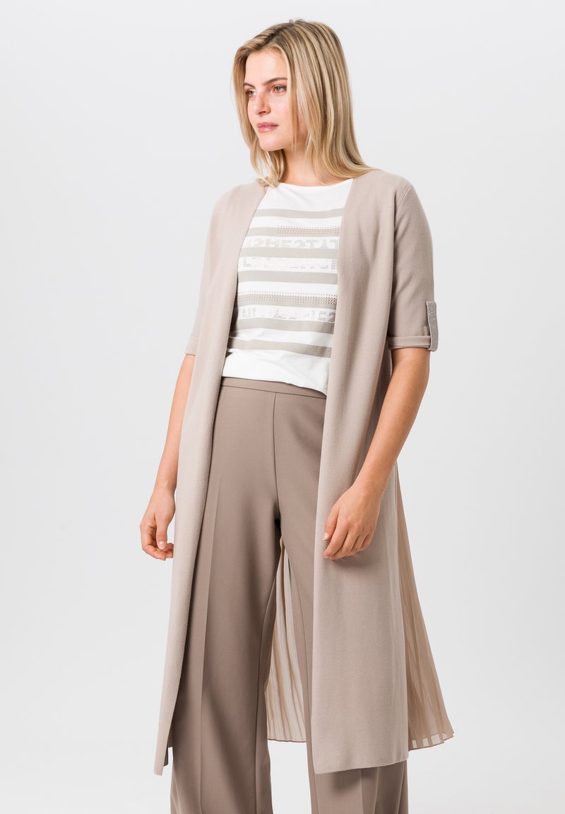 Cardigan long beige à manches courtes, au design drapé, texture lisse et dos plissé. Associé à un haut rayé et un pantalon beige.