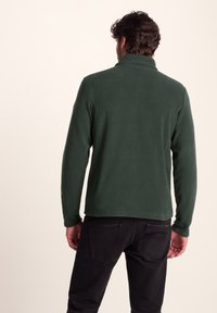 Dark green fleece jacket with a high collar, long sleeves, and a smooth texture, paired with black pants featuring back pockets.