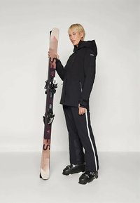 Svart skijakke med hette og glidelåslommer, parret med svarte skibukser med hvite striper. Modellen holder en ski med bindinger og et mønster.