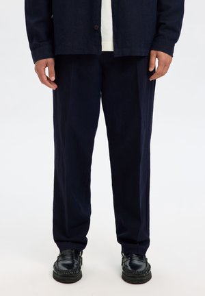 Chemise en lin bleu marine et pantalon large, associés à des mocassins en cuir noir. Le pantalon a une texture lisse, la chemise comporte une fermeture à un bouton.