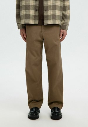 Personne portant un pantalon droit marron, des chaussures noires, un pull marron et une chemise à carreaux beige et marron, debout sur un fond blanc.