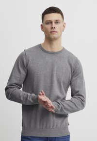 Grå strikket genser med rund hals og lange ermer, med teksturert stoff og ribbede mansjetter og linning. Modellen har hendene samlet.