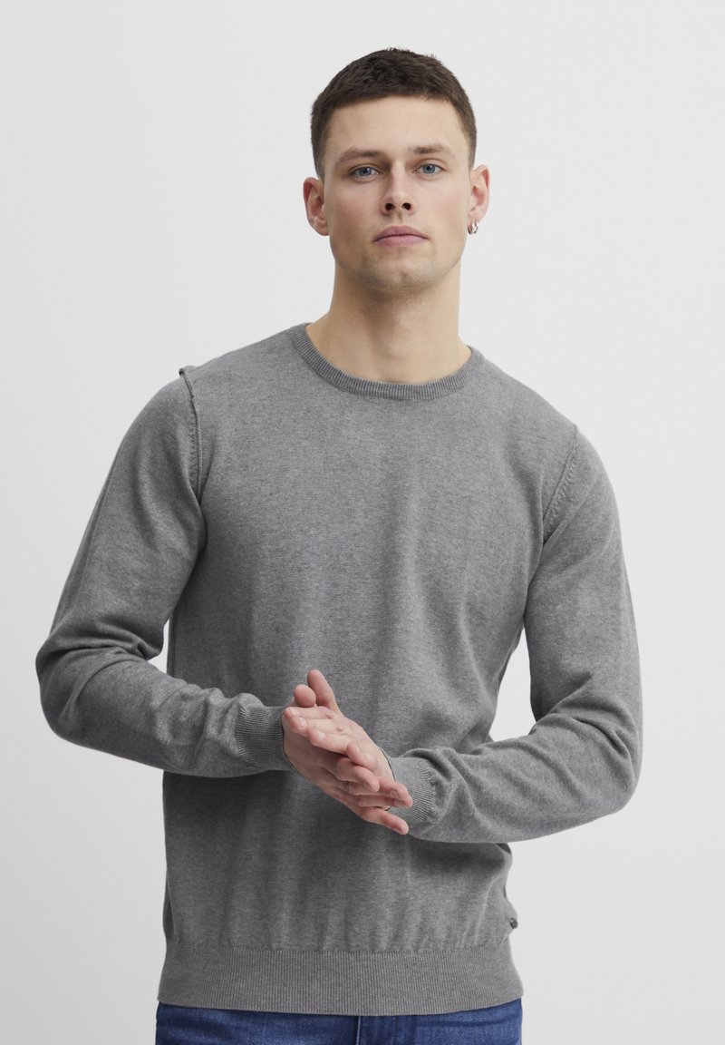Grå strikket genser med rund hals og lange ermer, med teksturert stoff og ribbede mansjetter og linning. Modellen har hendene samlet.