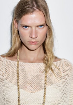 Lichtbeige gebreide trui met open geweven structuur, met een brede halslijn en gelaagd met een geborduurde ketting. Glad haar valt langs het gezicht.