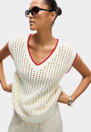 Top in maglia senza maniche a scollo a V di colore crema con dettagli rossi; presenta una texture a tessitura aperta e orlo a coste. Il modello posa con le mani sui fianchi.