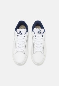 Baskets en cuir blanc avec un col en tissu bleu marine, dotées de lacets blancs et d'un logo près de la languette. Texture lisse et design minimaliste.