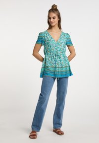 Ein türkisfarbener Blümchen-Peplum-Top mit V-Ausschnitt und kurzen Ärmeln, mit einem Kordelzug in der Taille und gemustertem Saum. Kombiniert mit blauen Jeans.