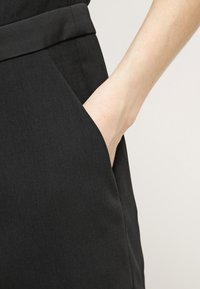 Main insérée dans la poche latérale d'un pantalon noir taille haute sur un fond clair uni.