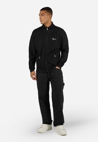 Sort ribbet jakkesæt med lynlås og høj krave, med hvid logo-broderi og forlommer, parret med sorte bukser og hvide sneakers.