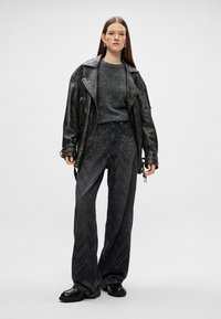Jaqueta de motoqueiro de couro preto sobre um suéter cinza, combinada com jeans pretos de perna larga com um padrão de ziguezague e um brilho sutil. Botas pretas.