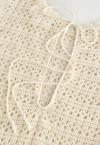 Top de encaje crema con un tejido abierto texturizado, escote con forma de ola y cierre de lazo en la parte posterior. Tela ligera y transpirable.