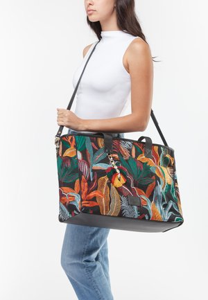 Femme portant un haut blanc sans manches et un jean bleu, portant un grand sac noir avec un motif de feuilles tropicales colorées et un pompon.