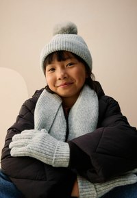 Gaiši zils adīts cepure ar pomponu, atbilstoša šalle un cimdi. Melna pildīta jaka, sēdoša poza uz neitrāla fona.